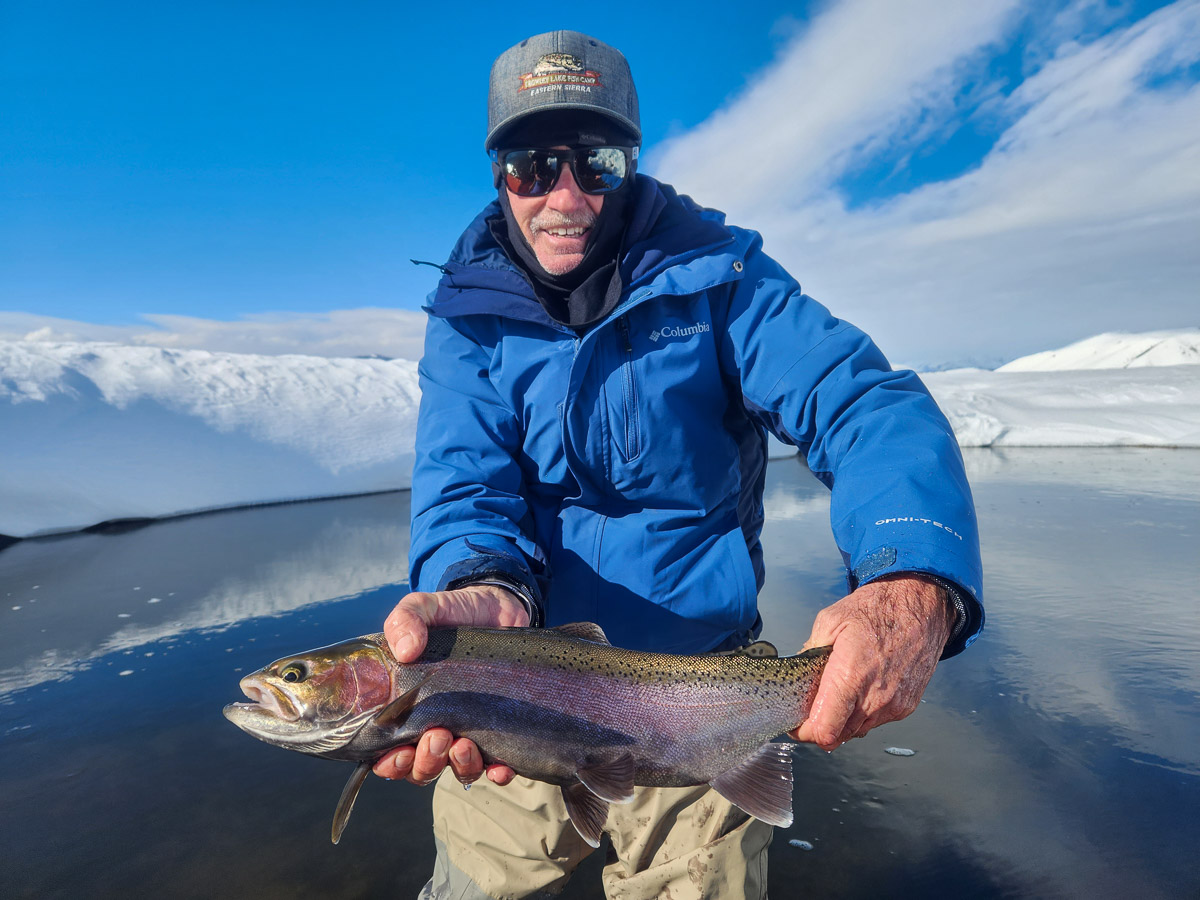 A smiling fisherman holding a massive rainbow trout next to the Upper Owens River in the snow in the eastern sierra fishing zone.