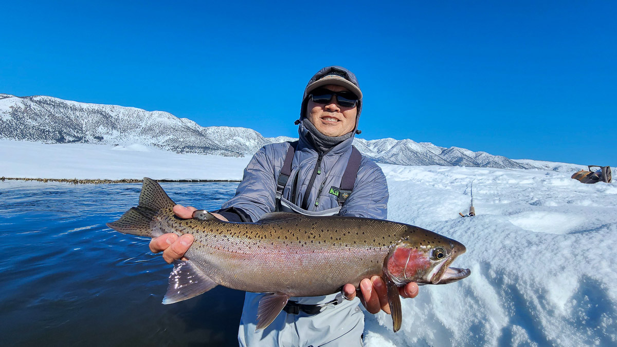 A smiling fisherman holding a massive rainbow trout next to a river in the snow on the Upper Owens River.