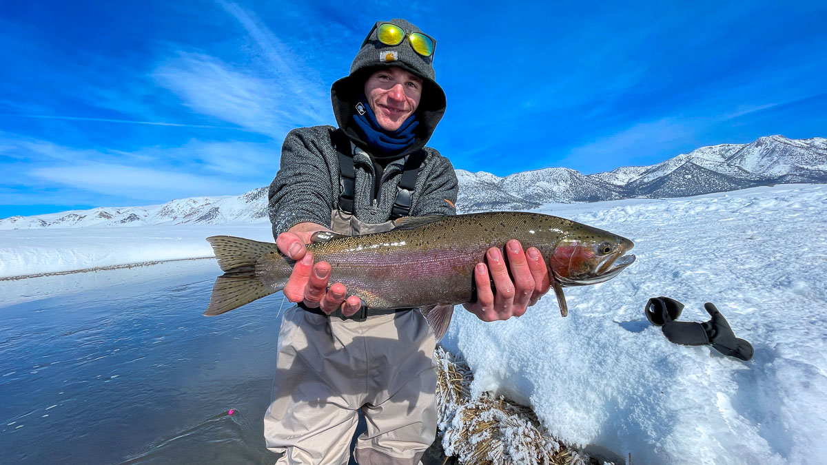 A smiling fisherman holding a rainbow trout in a river in the eastern sierra fishing zone.