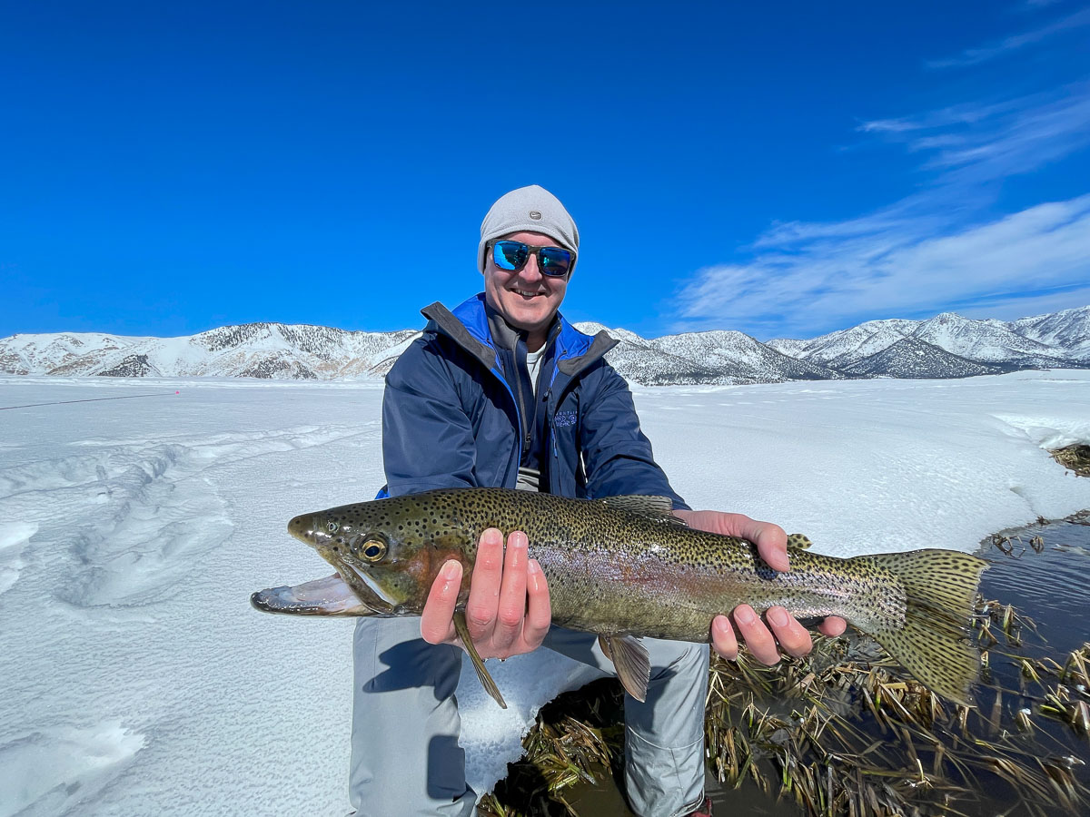 A smiling fisherman holding a massive rainbow trout next to a river in the snow in the eastern sierra fishing zone.