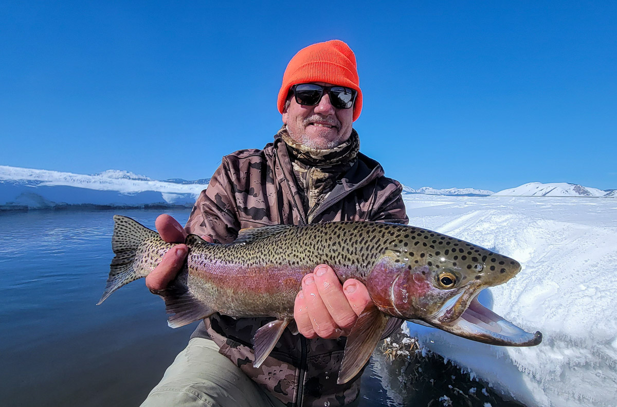 A smiling fisherman holding a massive rainbow trout next to the Upper Owens River in the snow in the eastern sierra fishing zone.