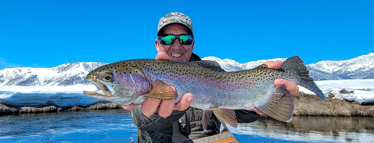 A smiling fisherman holding a massive rainbow trout next to the Upper Owens River in the snow in the eastern sierra,