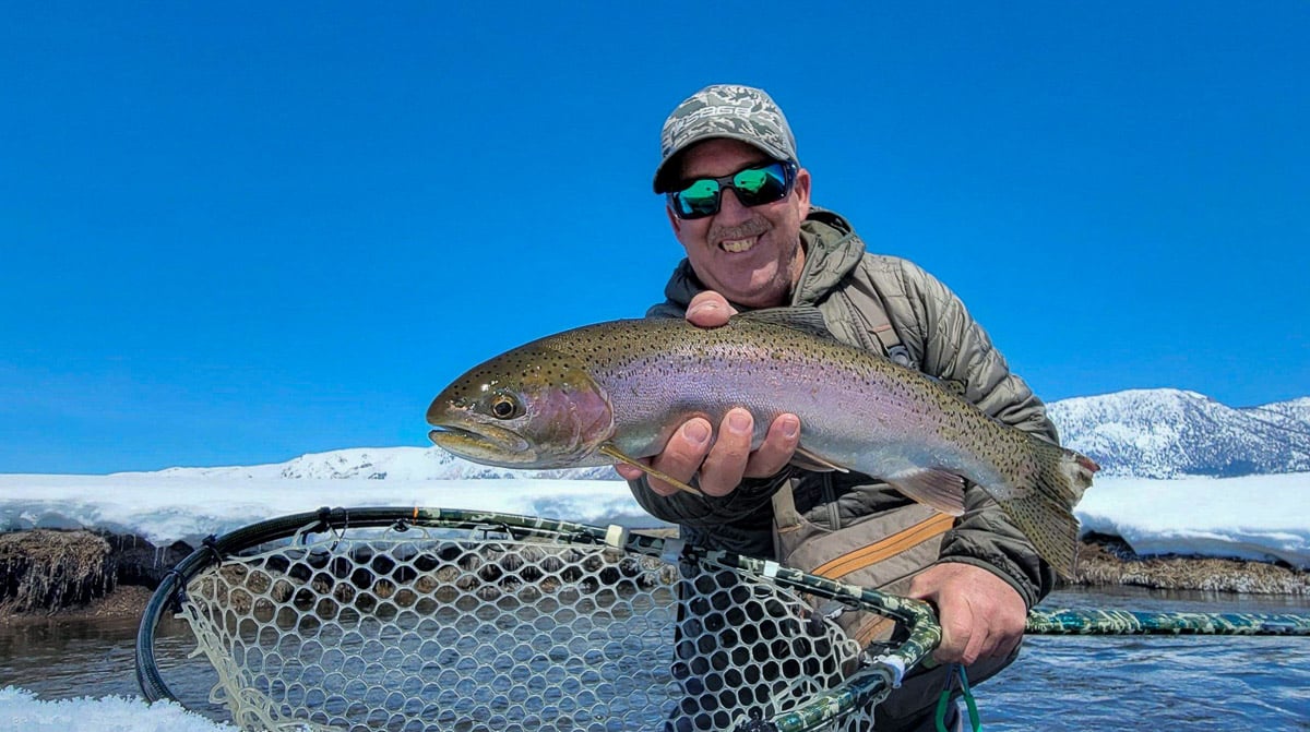 A smiling fisherman holding a massive rainbow trout next to a river in the snow on the Upper Owens River.