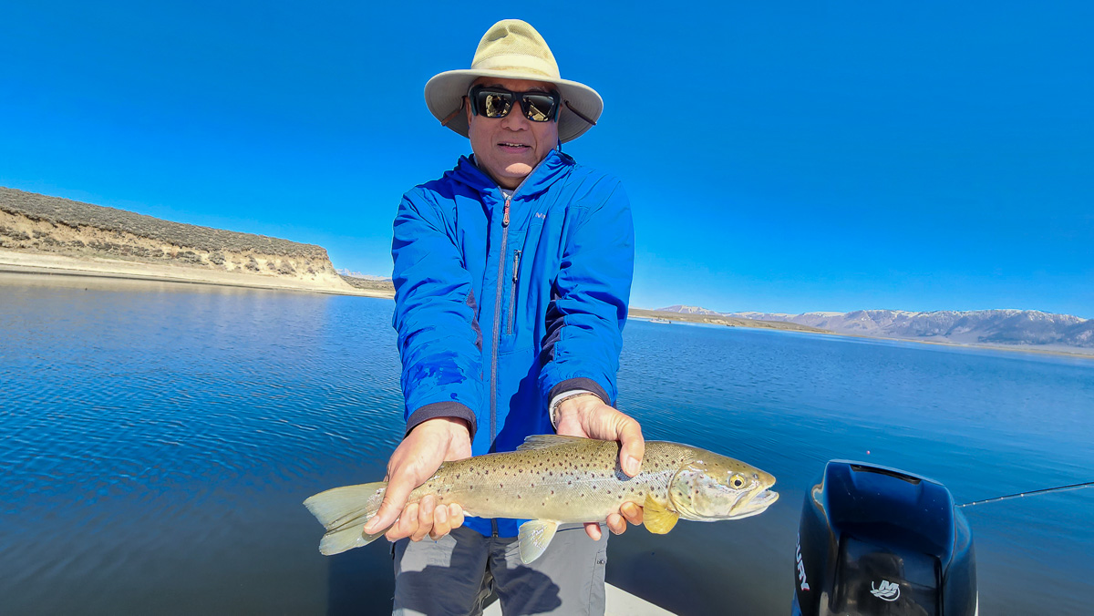 A fly fisherman on a boat on a lake with a large rainbow trout.