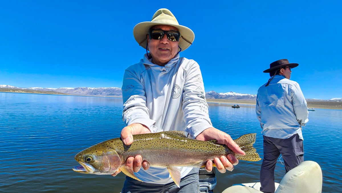 A fly fisherman on a boat on a lake with a large rainbow trout.