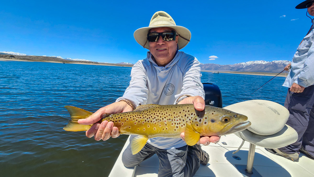 A fly fisherman on a boat on a lake with a large brown trout.