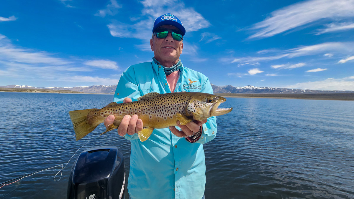 A fly fisherman on a boat on a lake with a large brown trout.