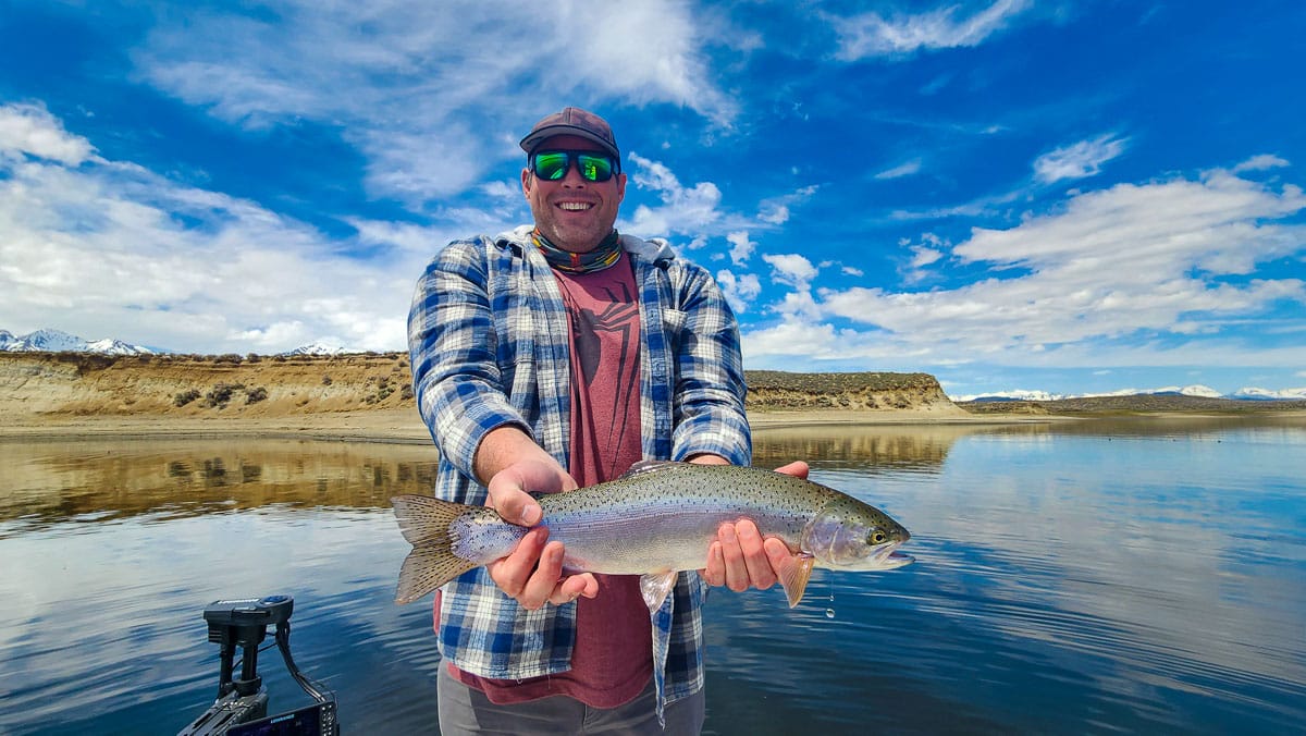 A fly fisherman on a boat on a lake with a large rainbow trout.