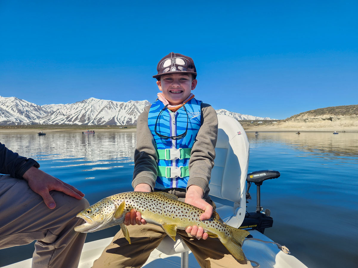A young fisherman holding a large brown trout from Crowley Lake on a boat.