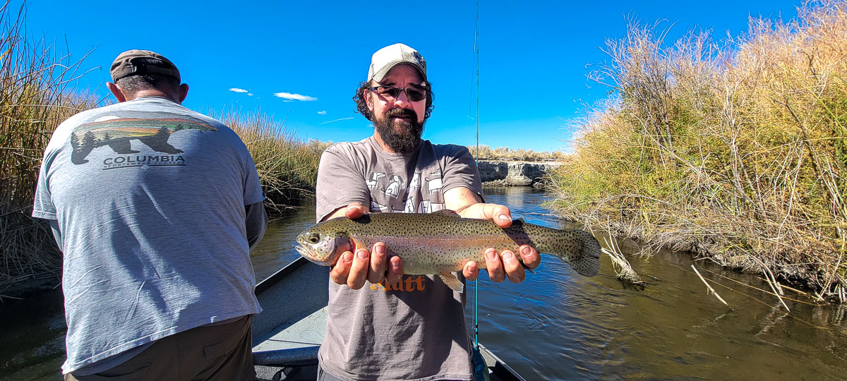 A smiling angler holding a large rainbow trout from a drift boat on the Lower Owens River.