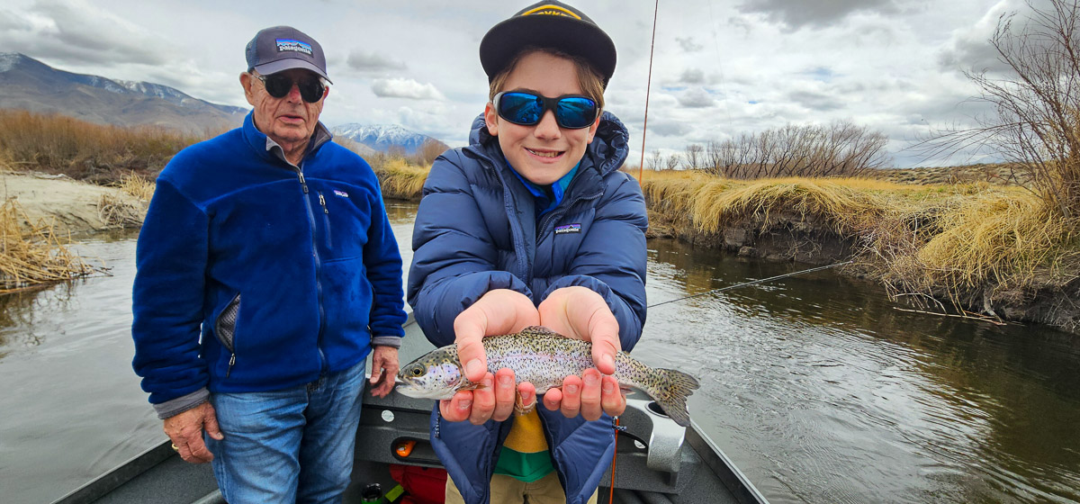 A young smiling fisherman with a rainbow trout in a drift boat on the Lower Owens River.