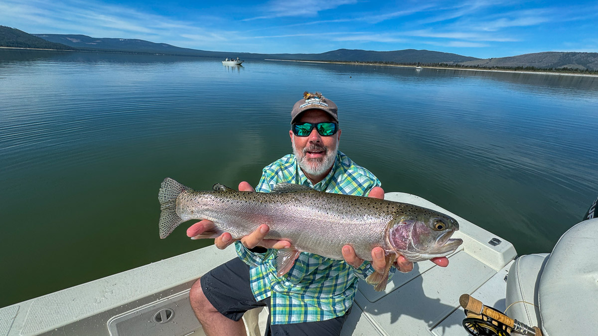 A hooded smiling fly fisherman holding a giant rainbow trout in a boat on Bridgeport Reservoir.