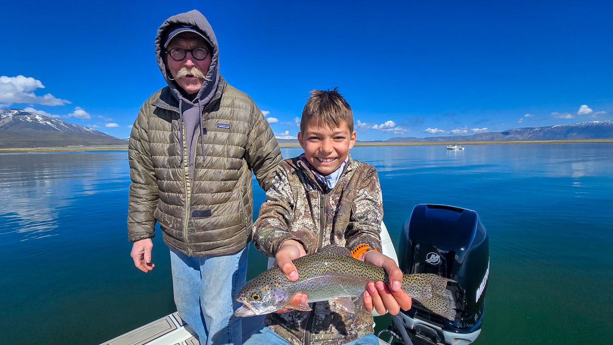 A young boy with a camouflage jacket holding a large rainbow trout next to an older man with glasses and a hood on Crowley Lake in a boat near Mammoth Lakes, CA