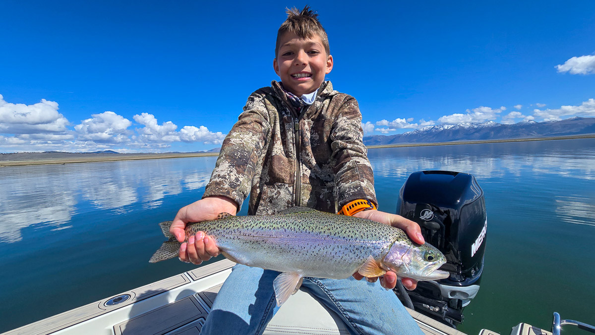 A young boy with a camouflage jacket holding a large rainbow trout on Crowley Lake in a boat near Mammoth Lakes, CA